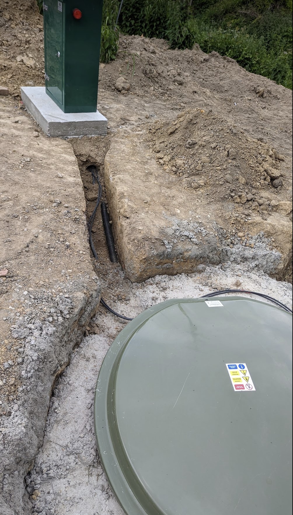 The green lid of the installed Klargester BioDisc, set into the ground with the control cabinet behind.